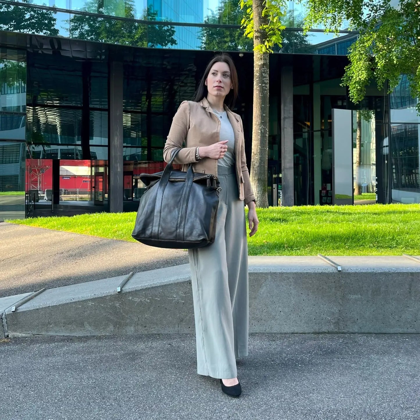 A woman in a neutral outfit carries the
brown leather Journey handbag by SUDO Zurich while standing outside a modern glass building.